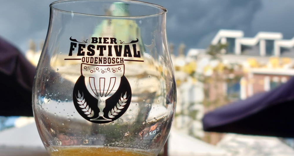 Een glas met het logo van het Bierfestival Oudenbosch, omringd door een sfeervolle festival locatie.