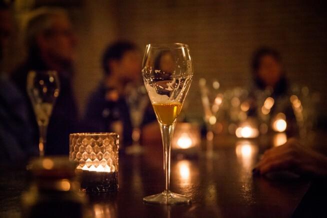 Glazen en kaarslicht op een houten tafel met mensen op de achtergrond in warme sfeer
