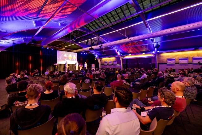 Een volle conferentiezaal met mensen die naar een presentatie op het podium kijken onder kleurrijke verlichting