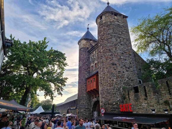 Mensen bij een historisch stenen gebouw tijdens een bierfestival met kraampjes en bomen.