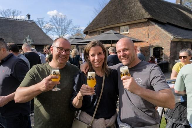 Drie mensen met glazen craft bier bij een festival met een rustieke achtergrond.