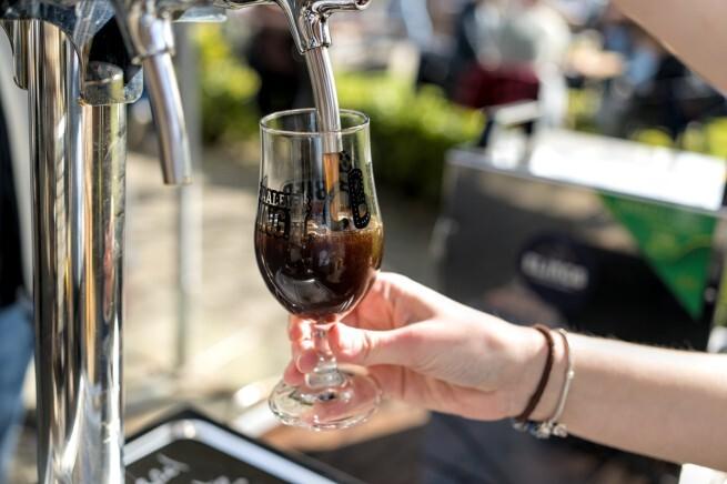 Een persoon tapt een donker craft bier in een helder glas op een zonnig festival.
