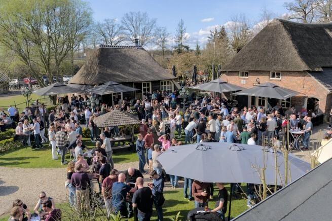 Mensen genieten van een craft bier festival buiten in de zon met rieten daken op de achtergrond.