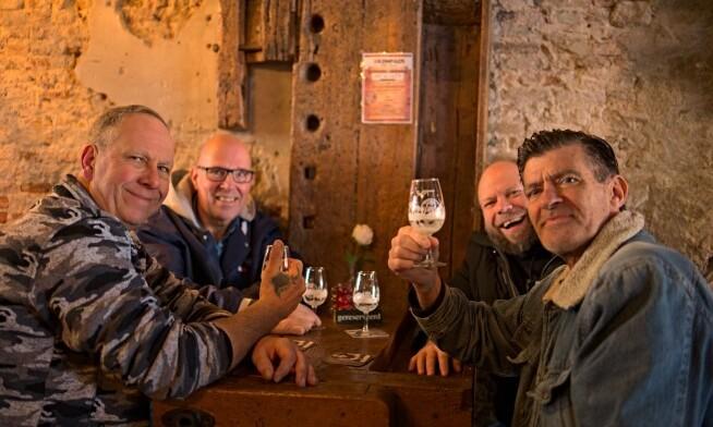 Vier mannen genieten van een biertje in een gezellige, rustieke bar met stenen muren.