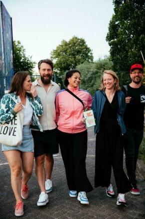 Vijf lachende mensen lopen buiten in informele kleding op een zonnige dag.