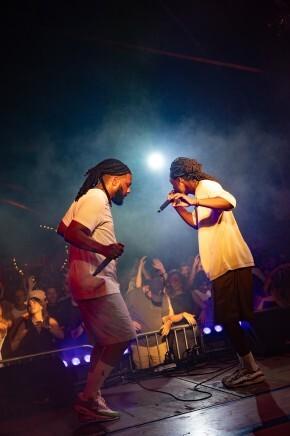 Twee artiesten op een podium met microfoons tijdens een optreden met publiek op de achtergrond.