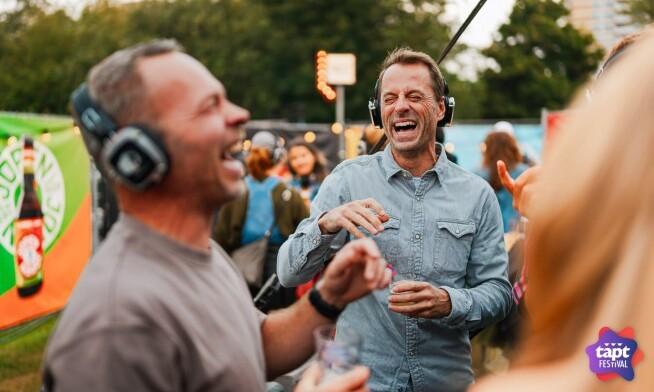 Twee mannen lachen en dragen koptelefoons tijdens een buitenfestival met kleurrijke achtergronden.