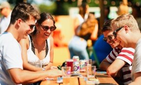 Groep vrienden genietend van drankjes aan een houten tafel op een buitenfestival