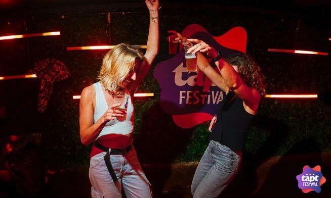 Twee mensen dansen met drankjes tijdens een festival met kleurrijke verlichting.