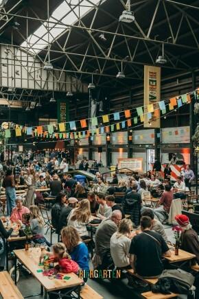 Drukke hal met tafels, kleurrijke vlaggen en mensen bij het Biergarten Lentefestival.