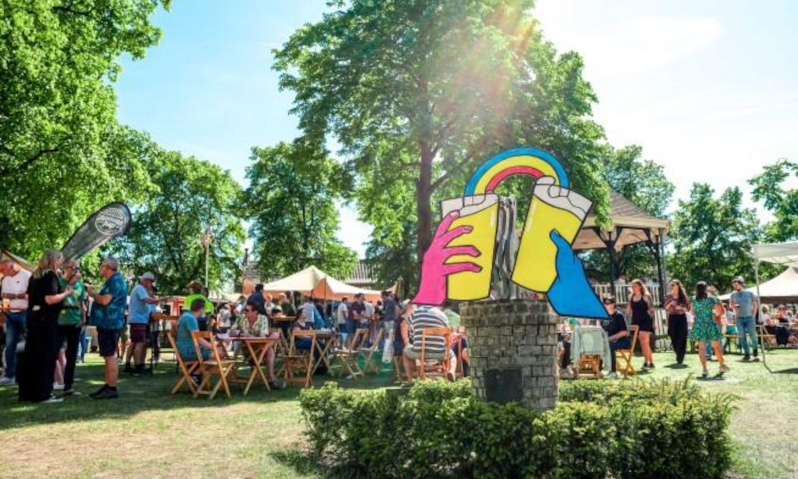 Mensen genieten van bierfestival in het park met kleurrijke decoraties en zonnige blauwe lucht.
