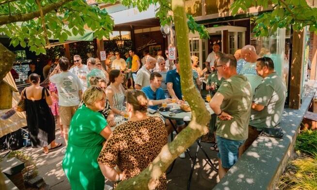 Groep mensen bij een buitenbijeenkomst onder groene bomen, pratend en genietend van drankjes.