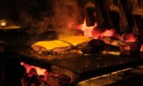 Hamburgers met kaas sissend op een grill boven houtskoolvuur in een donkere omgeving.