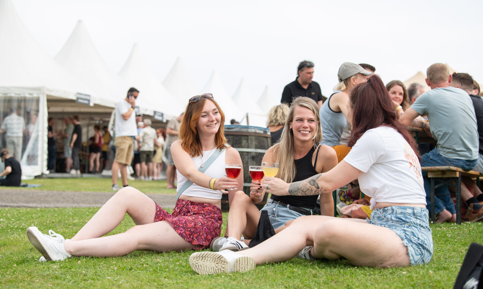Drie vrouwen zitten op gras en proosten met bier tijdens festival met witte tenten op de achtergrond.