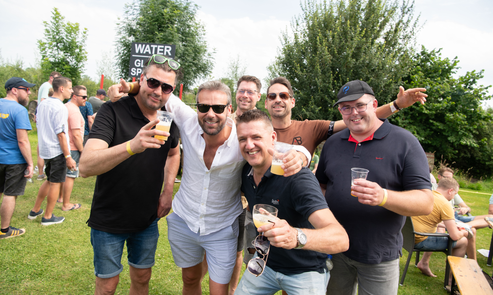 Groep vrienden glimlacht en proost met bier op een zonnig festival terwijl andere bezoekers ontspannen op het gras.