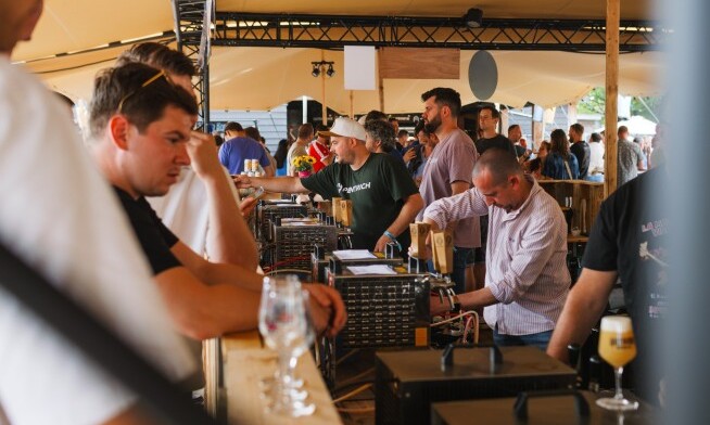 Mensen bij een bar met bierpompen tijdens een buitenfestival, onder een tentdak.