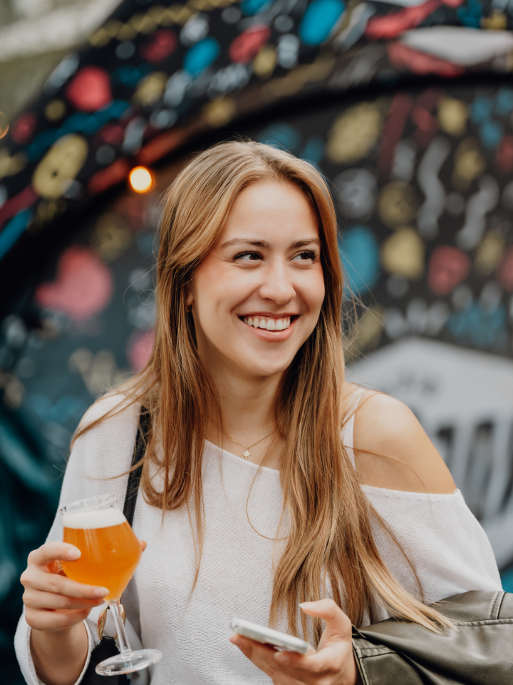 Vrouw met bierglas en smartphone lacht voor kleurrijke achtergrond.