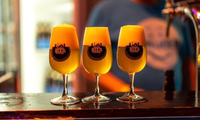 Drie glazen bier met logo van Helders Bier Festival op een bar bij avondlicht.