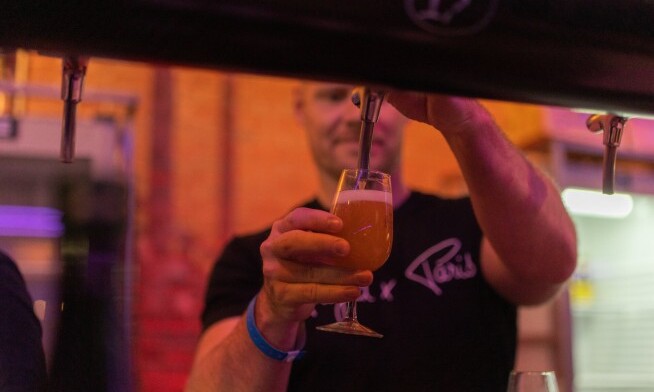 Persoon schenkt een glas bier in op een festival met warme achtergrondkleuren.