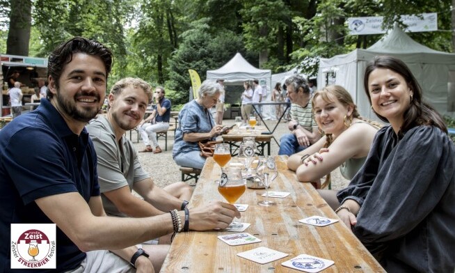 Groep mensen aan een houten tafel met bier bij Zeister Streekbierfestival in een parkachtige omgeving.