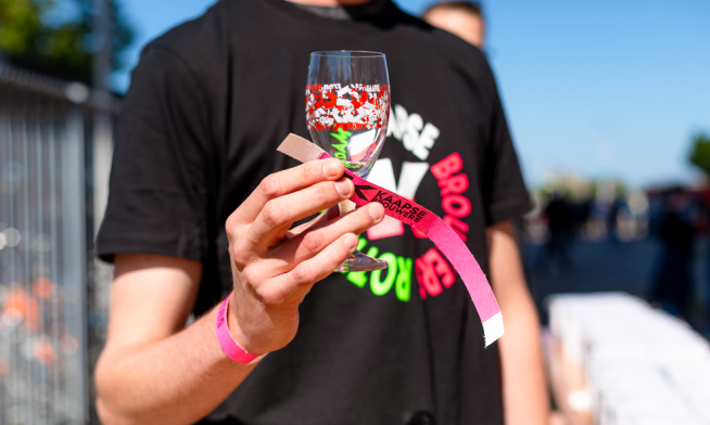 Persoon in zwart T-shirt houdt wijnglas met festivalbandje vast.