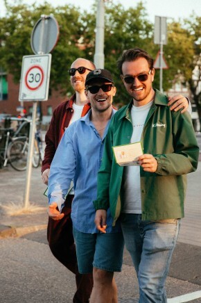 Drie lachende mannen wandelen buiten tijdens het avondlicht met een verkeersbord op de achtergrond.