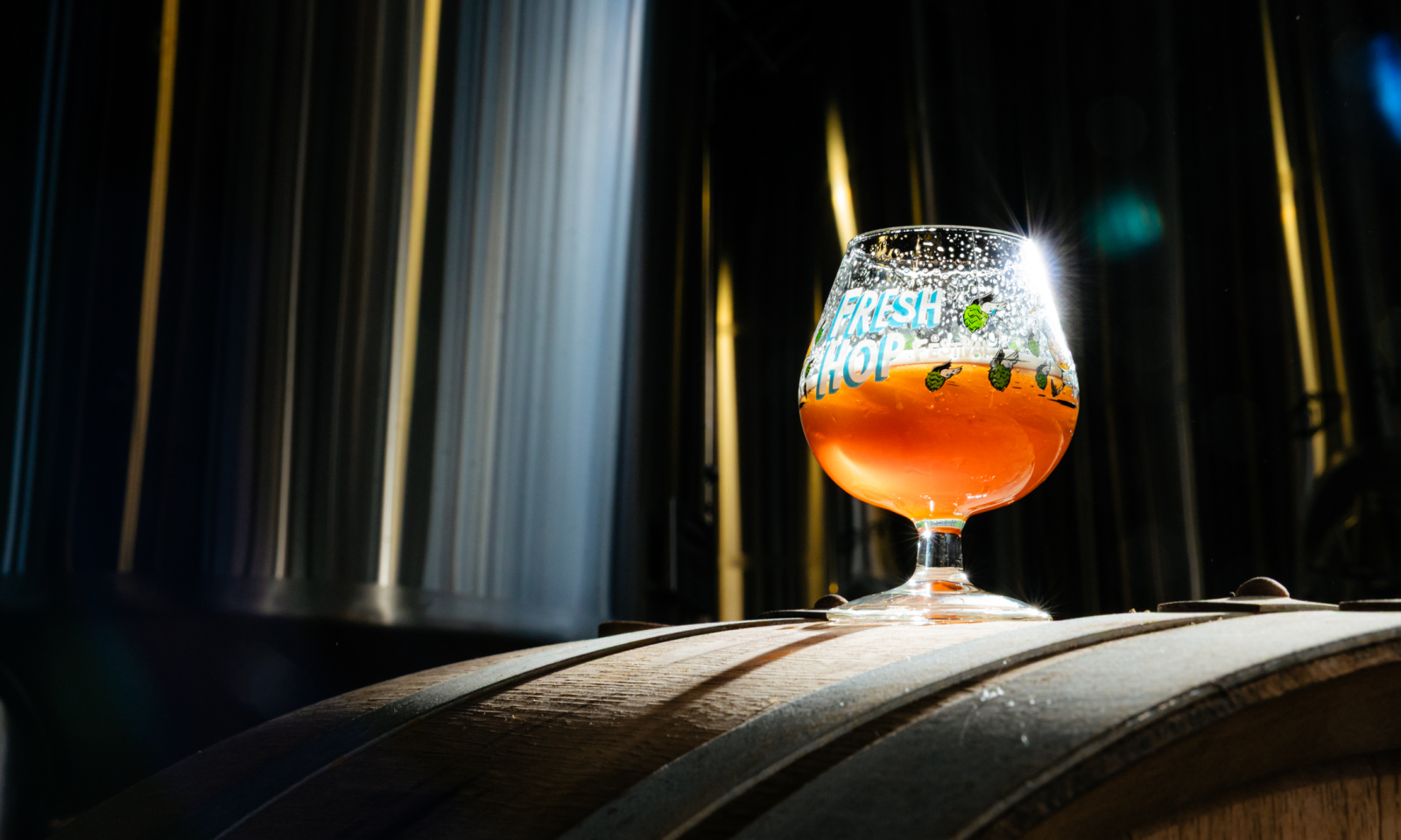 Glas amberkleurig Fresh hop-bier op een houten vat in een donkere brouwerij met metalen tanks.