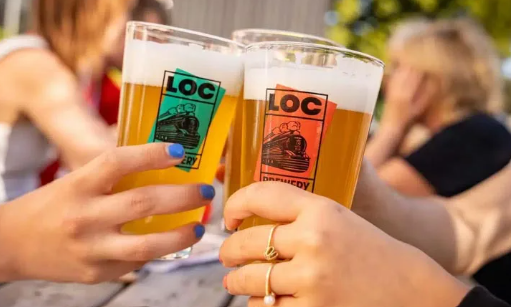 Twee handen proosten met plastic bekers bier op een zonnig terras met onscherpe mensen op de achtergrond.