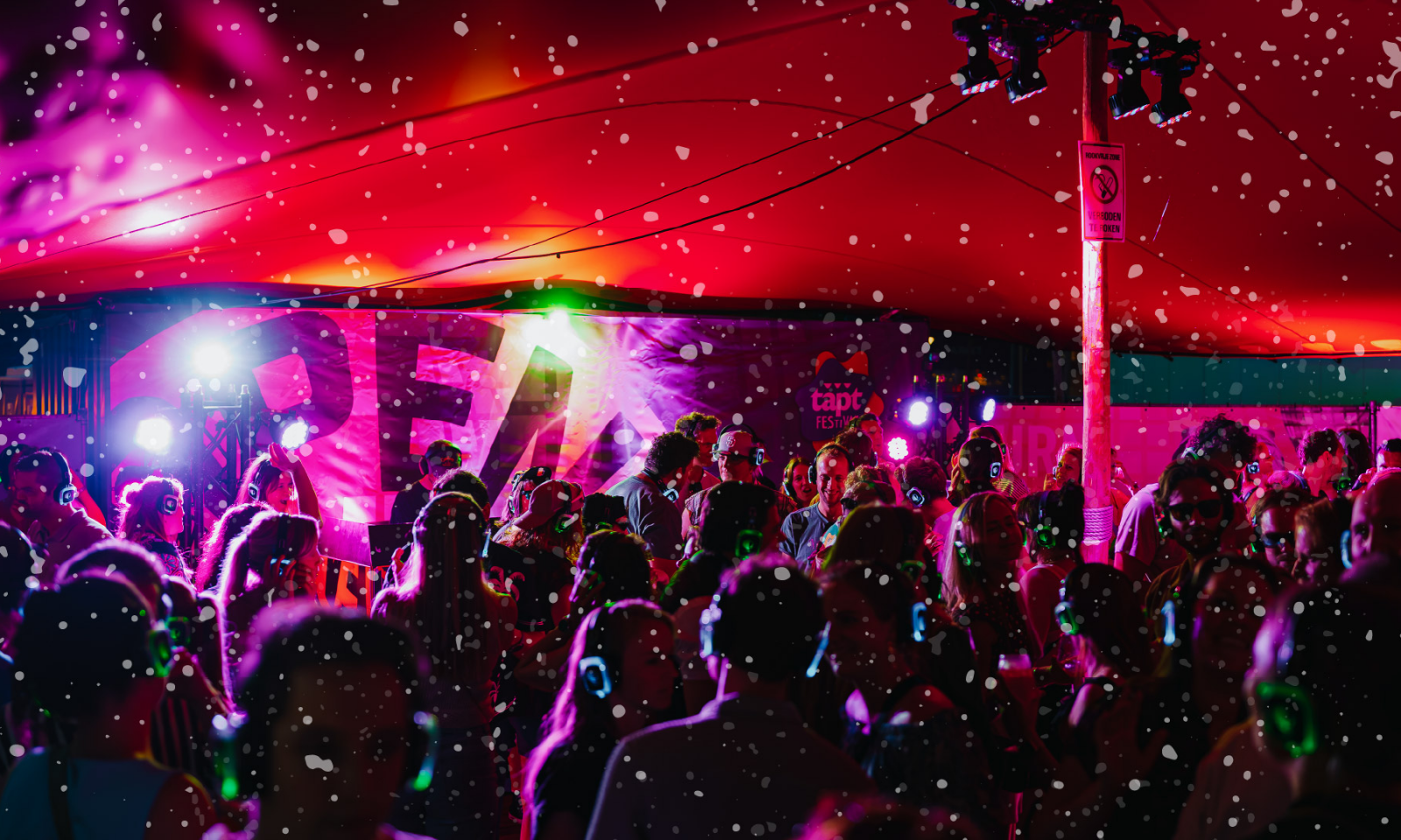 Feestende menigte met koptelefoons onder rode festivaltent met kleurrijke lichten en sneeuwvlokken.