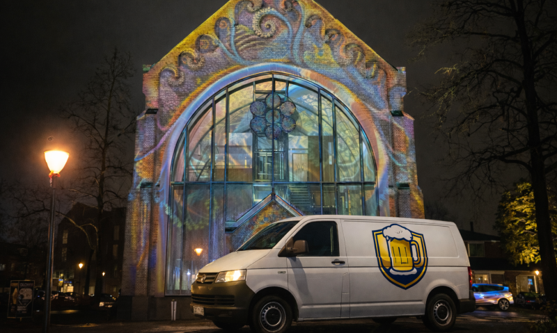 Wit bezorgbusje met een neutraal bierlogo zonder tekst, geparkeerd in de avond voor een historisch kerkgebouw dat verlicht is met kleurrijke lichtprojecties.