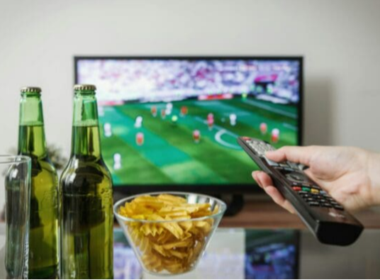 Twee bierflessen en een schaaltje met chips voor een tv met een voetbalwedstrijd.