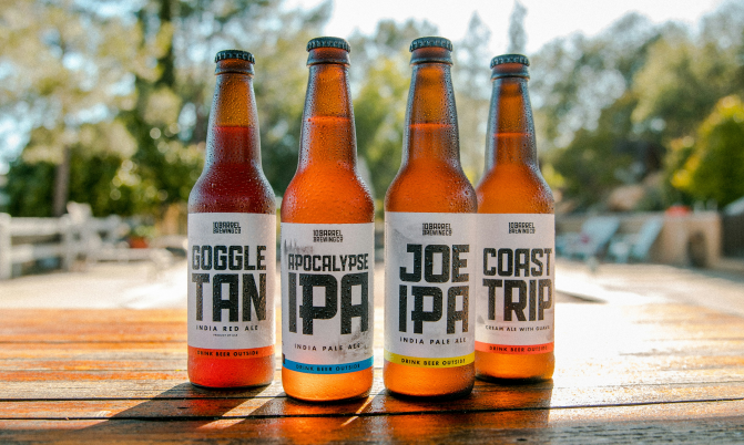 Vier soorten ambachtelijk bier in flessen staan op een houten tafel buiten, perfect voor een zonnige dag.