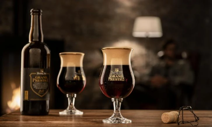 Twee glazen met donker bier op een tafel, naast een fles en een open haard op de achtergrond.