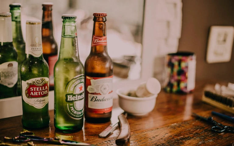 Drie bierflessen van verschillende merken op een houten tafel met scherpe voorwerpen naast een spiegel.