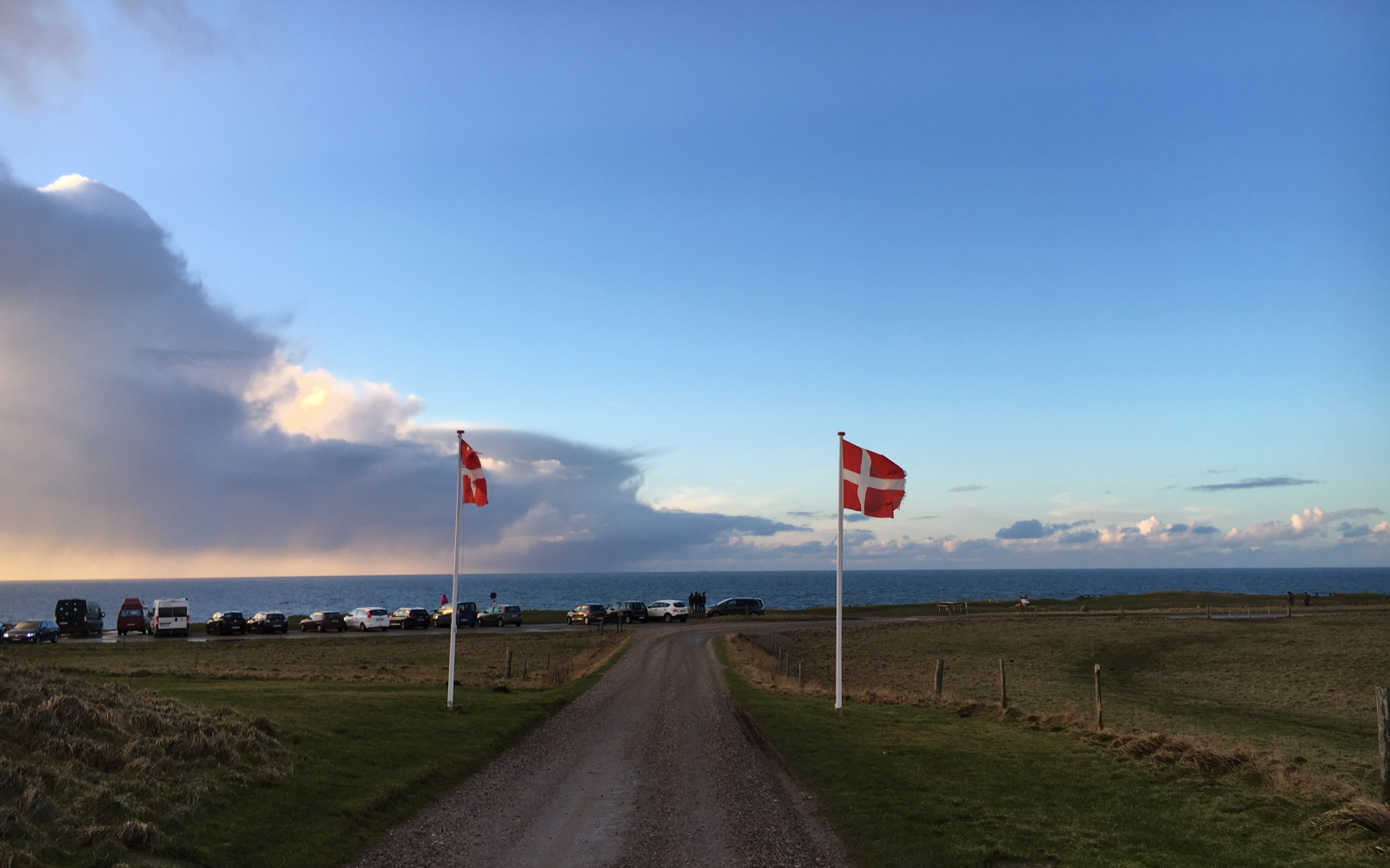 Deense vlaggen langs een onverharde weg, met een zicht op geparkeerde auto's en de zee in de verte.