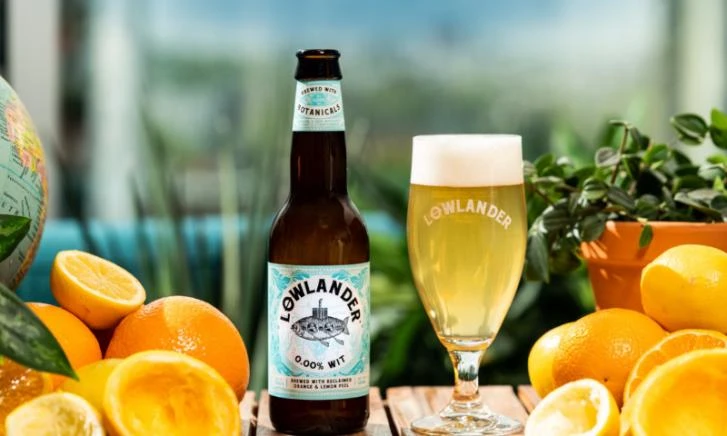 Fles en glas Lowlander 0.0% Witbier omringd door sinaasappels en planten.