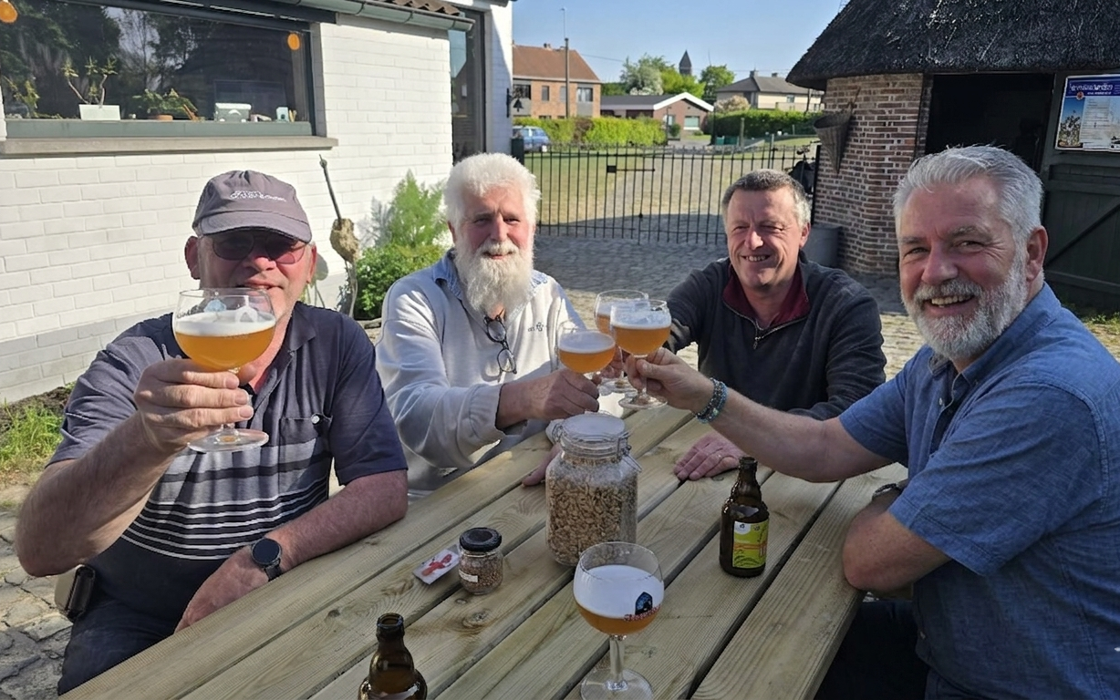 Vier mannen klinken met bier op een houten tafel buiten bij een wit gebouw.