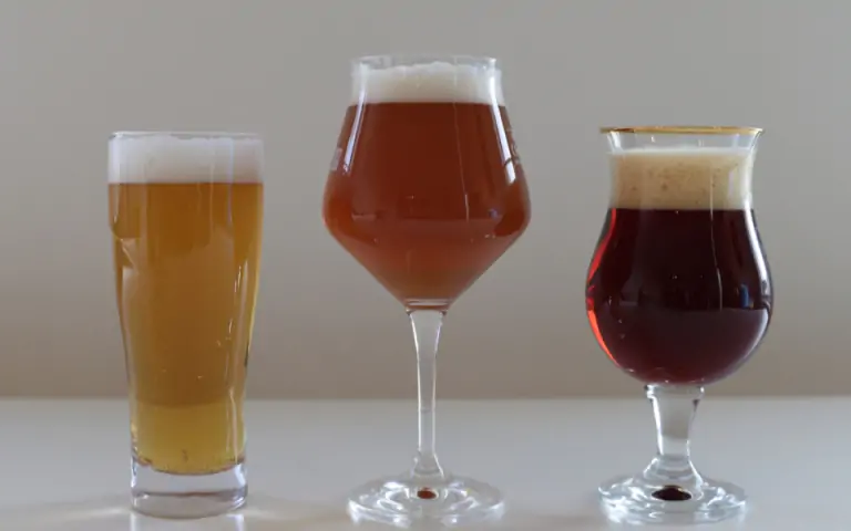 Drie verschillende glazen bier met schuimkraag op een witte tafel tegen een grijze achtergrond.