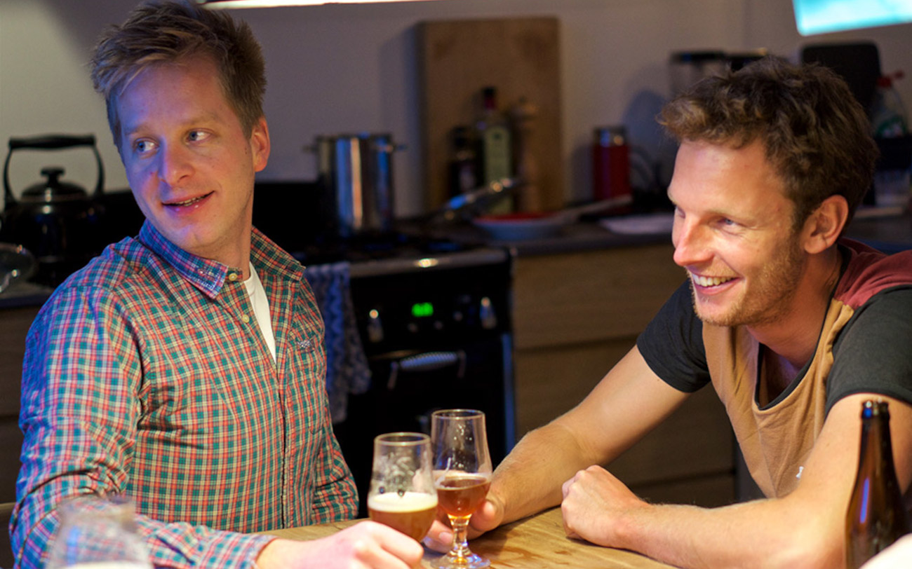 Twee mannen zitten aan een keukentafel met glazen bier, lachend en in gesprek