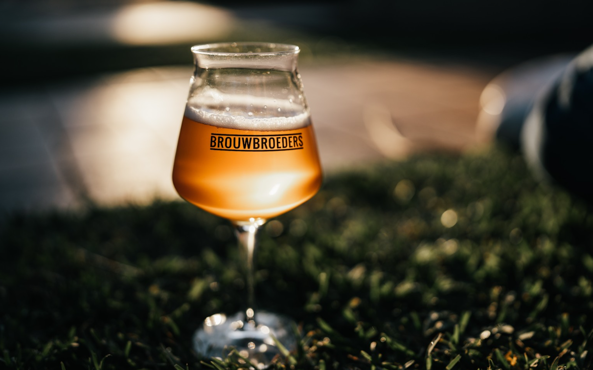 Glazen bier met 'Brouwbroeders'-logo op gras in avondlicht
