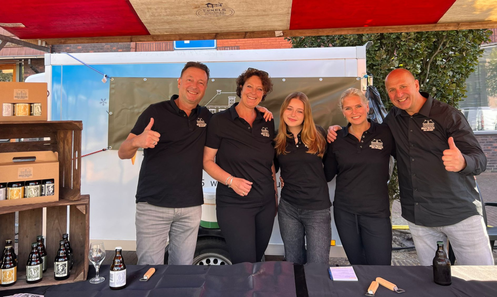 Groep mensen in zwarte polo's staan voor een kraam met bierflesjes en glazen.