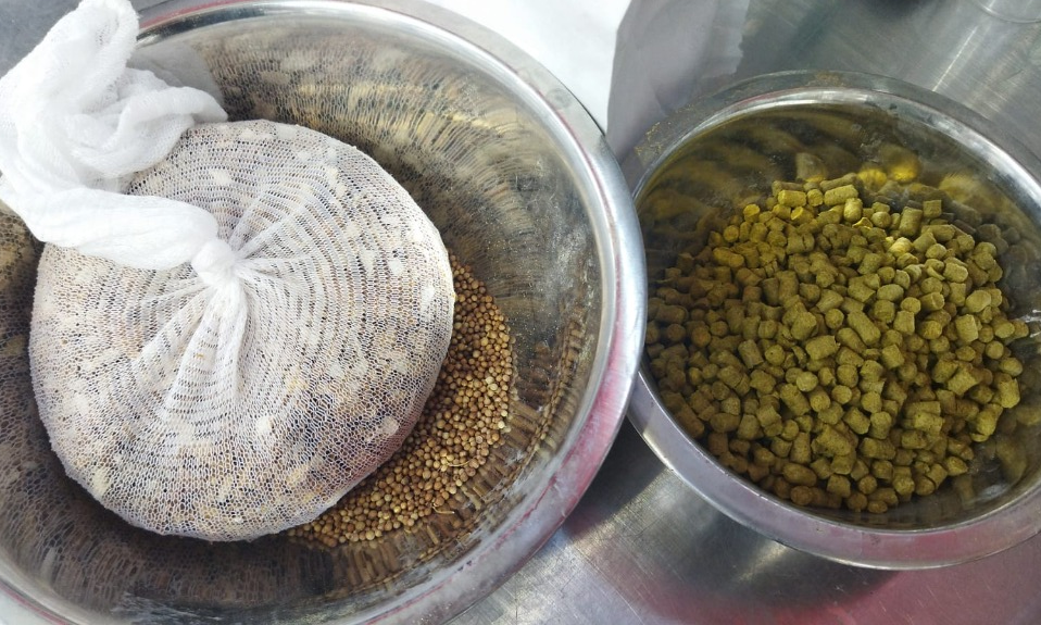 Twee metalen schalen met mout in een netzak en hopkorrels op een metalen tafel.