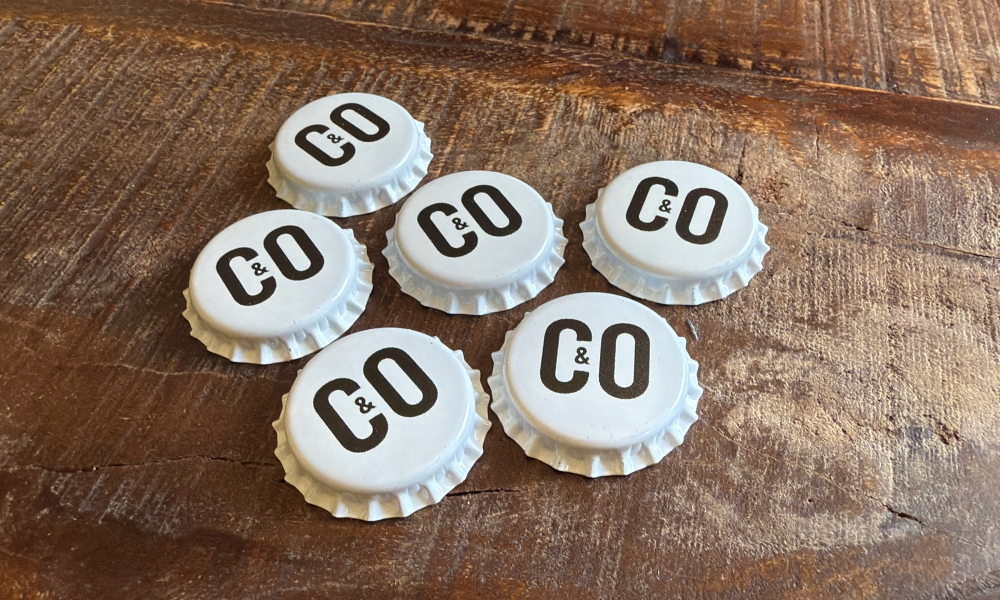 Zes witte flesdoppen met CO&CO logo op een houten tafel.
