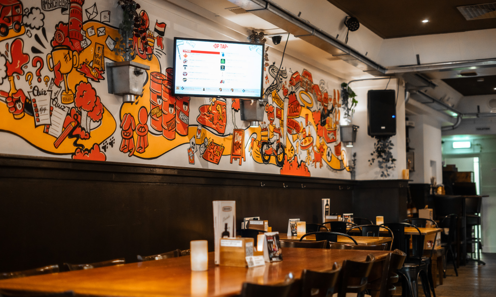Binnenkant van een gezellige bar met houten tafels en kleurrijke muurtekening.