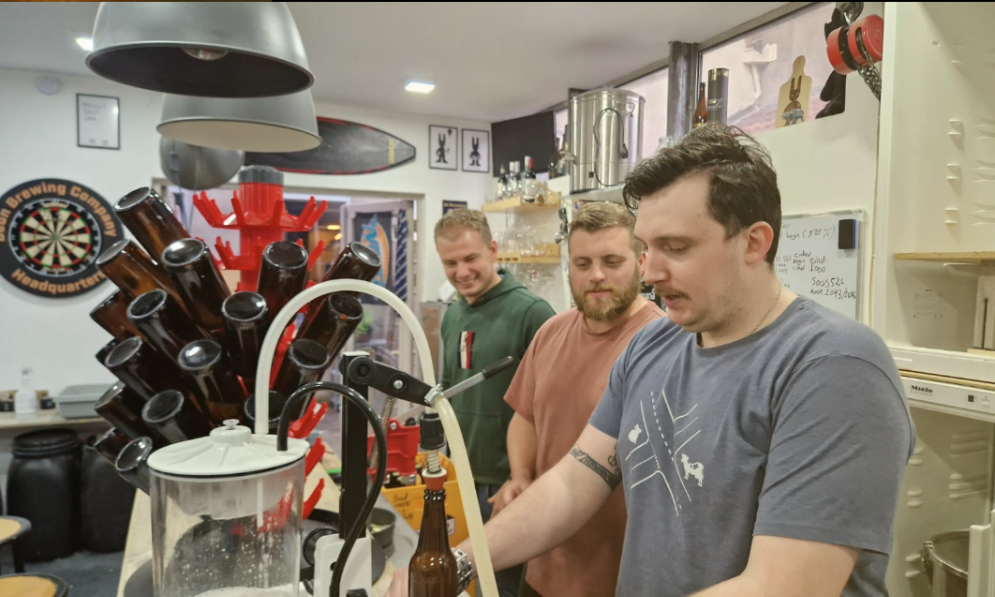 Drie mannen in een brouwerij met bierflessen en apparatuur.