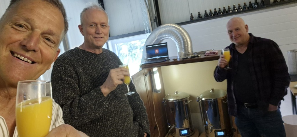 Drie mannen proosten met geel drankje in een ruimte met brouwketels en flessen op de achtergrond.