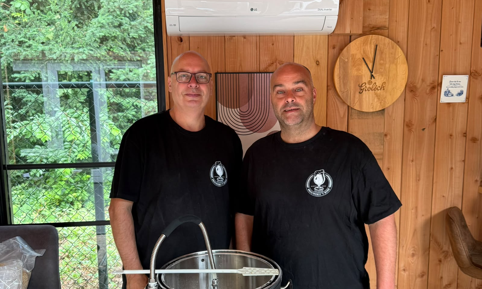 Twee mannen in zwarte shirts staan achter een brouwketel in een houten ruimte.