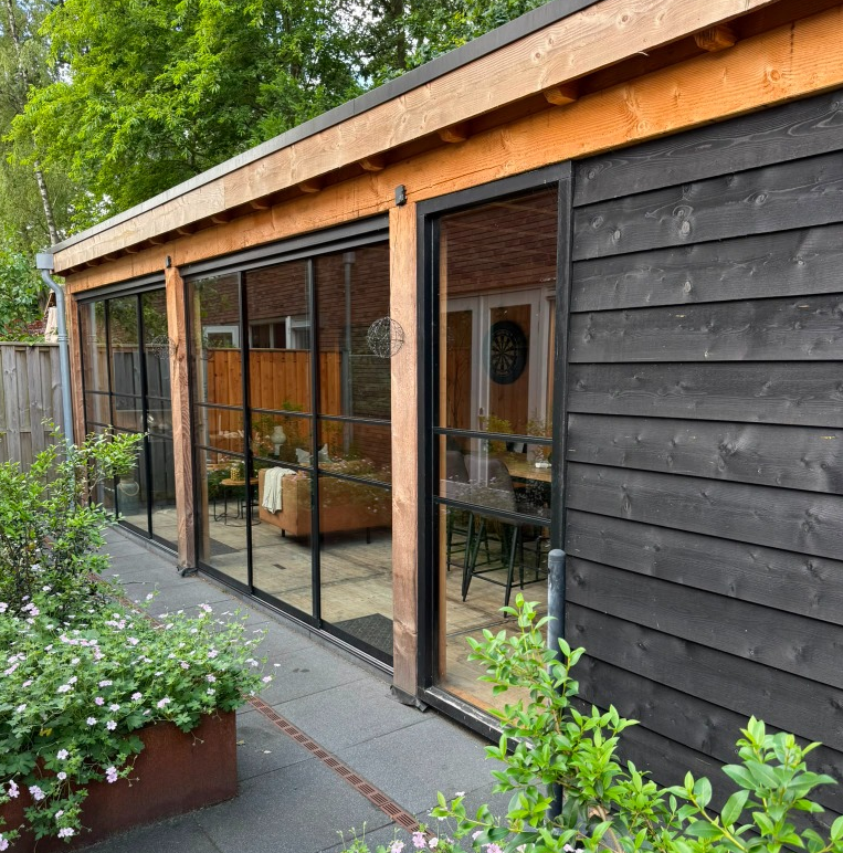 Glazen pui met zwarte kozijnen aan een houten tuinhuis, omringd door groene planten.