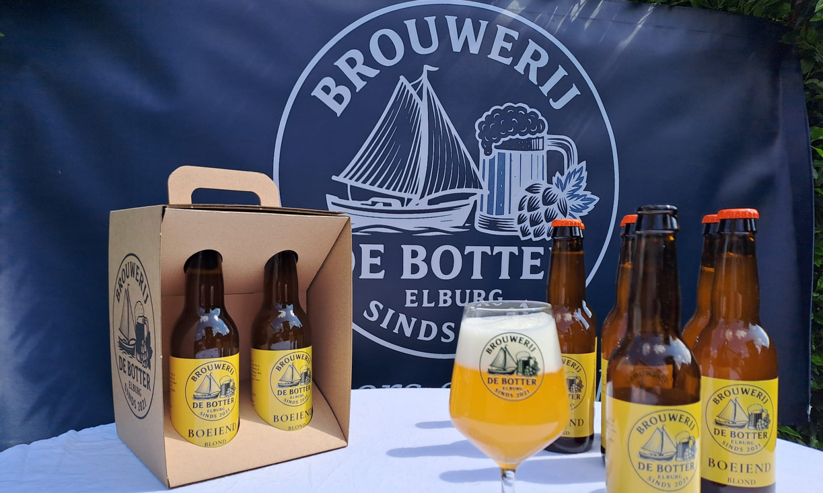 Bierflessen, glas en draagdoos van Brouwerij De Botter Boeiend blond voor een donkerblauwe banner.