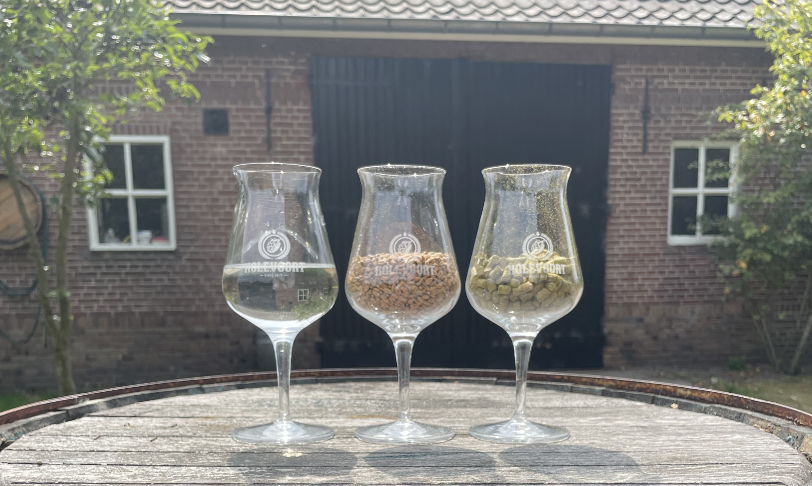Drie glazen met water, granen en hop op een houten vat voor een bakstenen schuur.